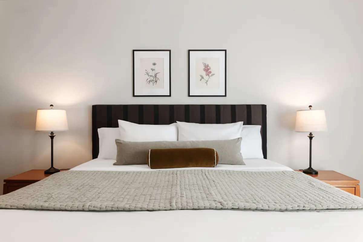 Bedroom with a king-size bed with a dark striped headboard, white and gray bedding, two lamps and two framed botanical prints.