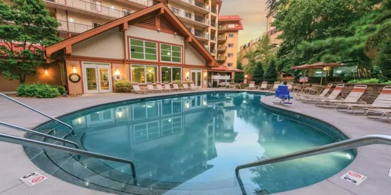 Outdoor pool with loungers in front of resort buildings with a wooden lodge-style roof.