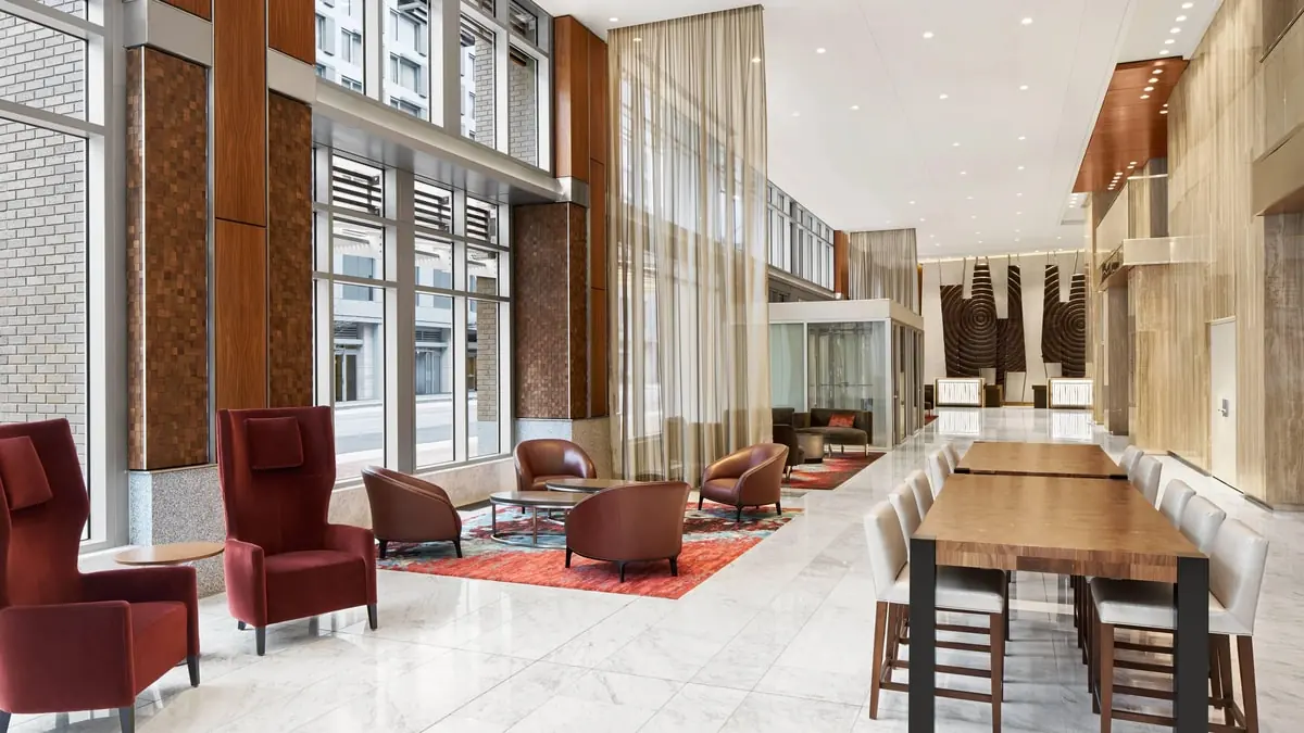Hotel lobby with tall windows, seating areas with red armchairs and a long wooden table with chairs.