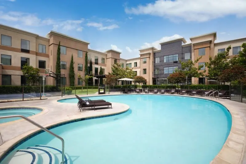 Curved outdoor swimming pool with loungers in front of hotel buildings.