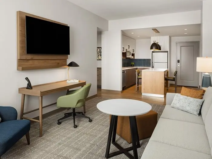 Living area with TV, desk, round table and sofa, kitchen behind.